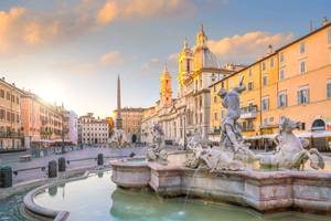 Twilight view of a white marble fountain with statues of horses and Neptune fighting an octopus with the baroque Piazza Navona square behind it