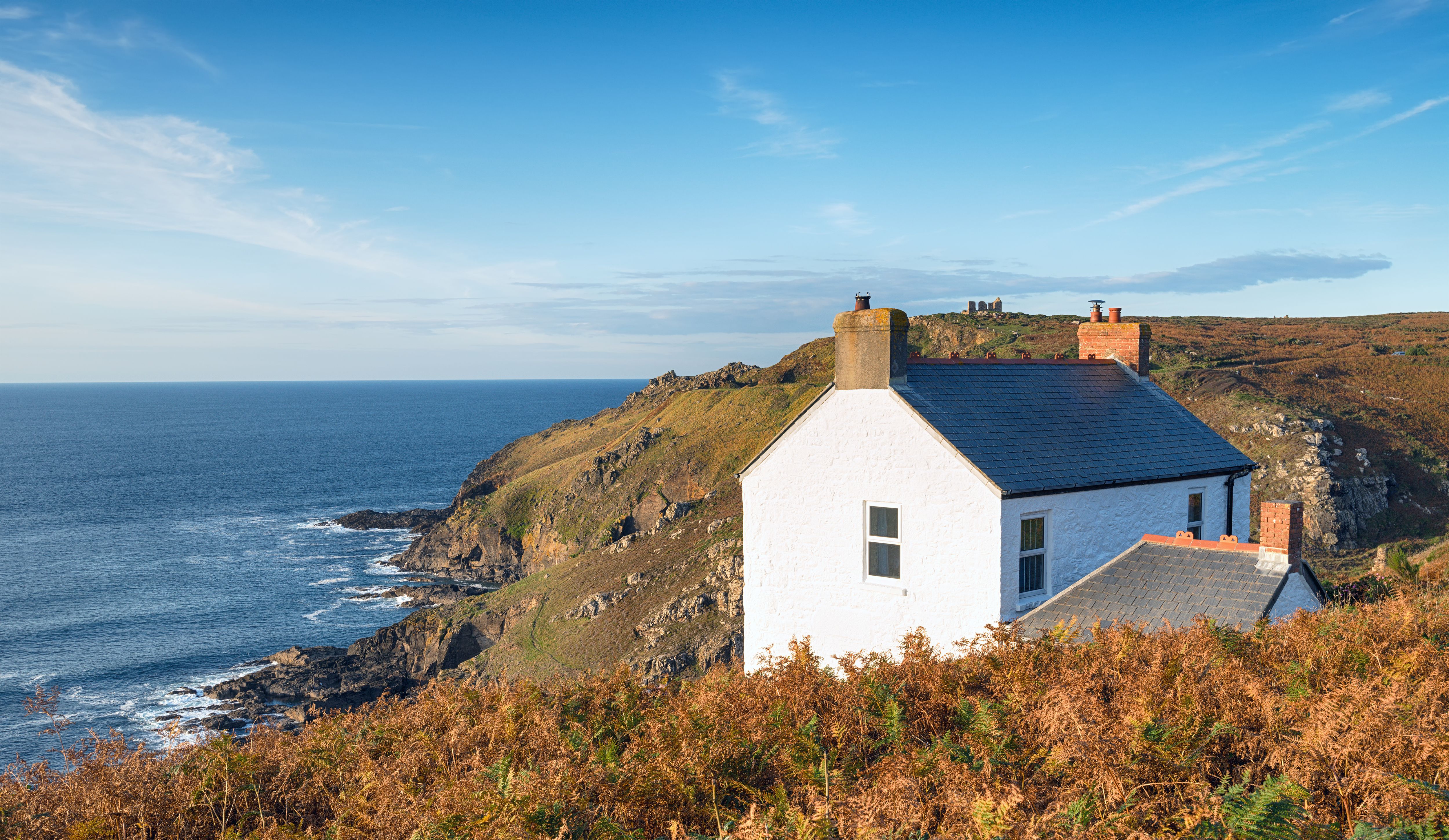 A whitewashed cottage perched on the cliffs of Cape Cornwall surrounded by autumnal brown and orange shrubs and looking out to the sea