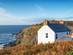 A whitewashed cottage perched on the cliffs of Cape Cornwall surrounded by autumnal brown and orange shrubs and looking out to the sea
