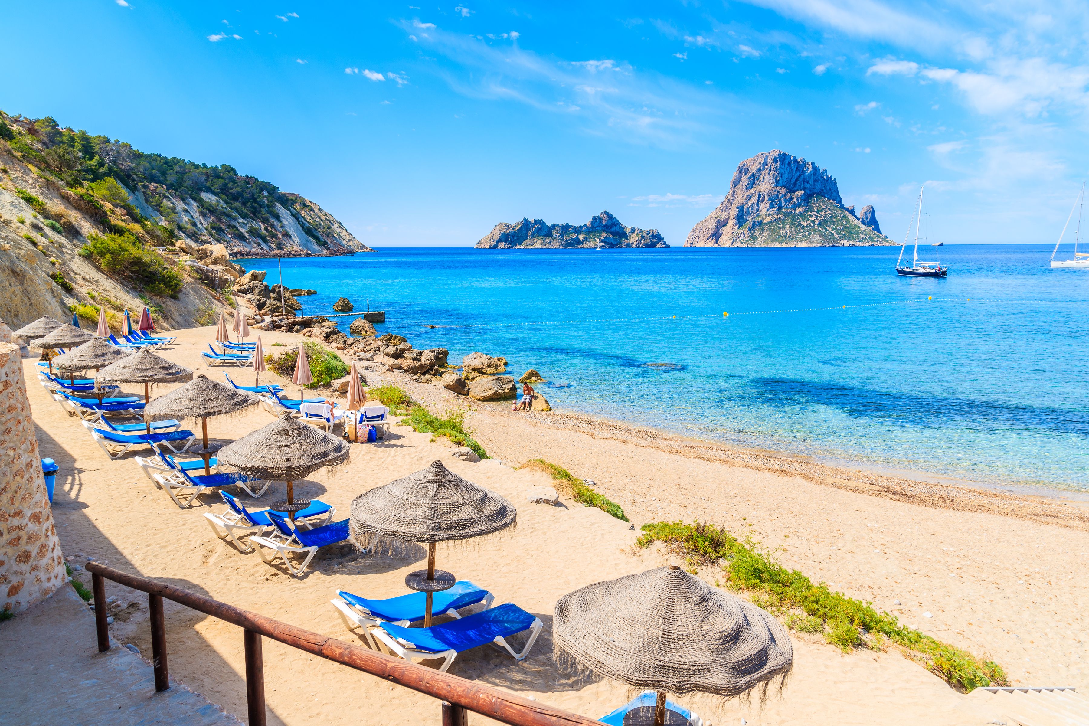 View of empty blue sun-loungers and thatched umbrellas on a golden beach that overlooks an imposing rock formation jutting out of the water