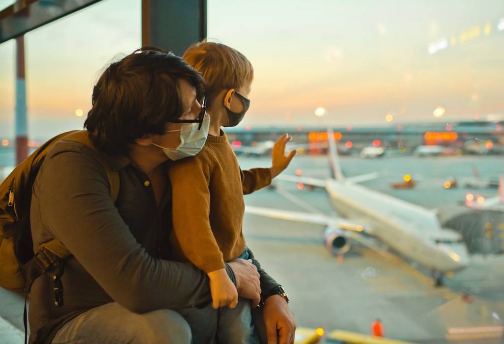 A father holding his son and looking out of an airport window both wearing protective coronavirus facemasks