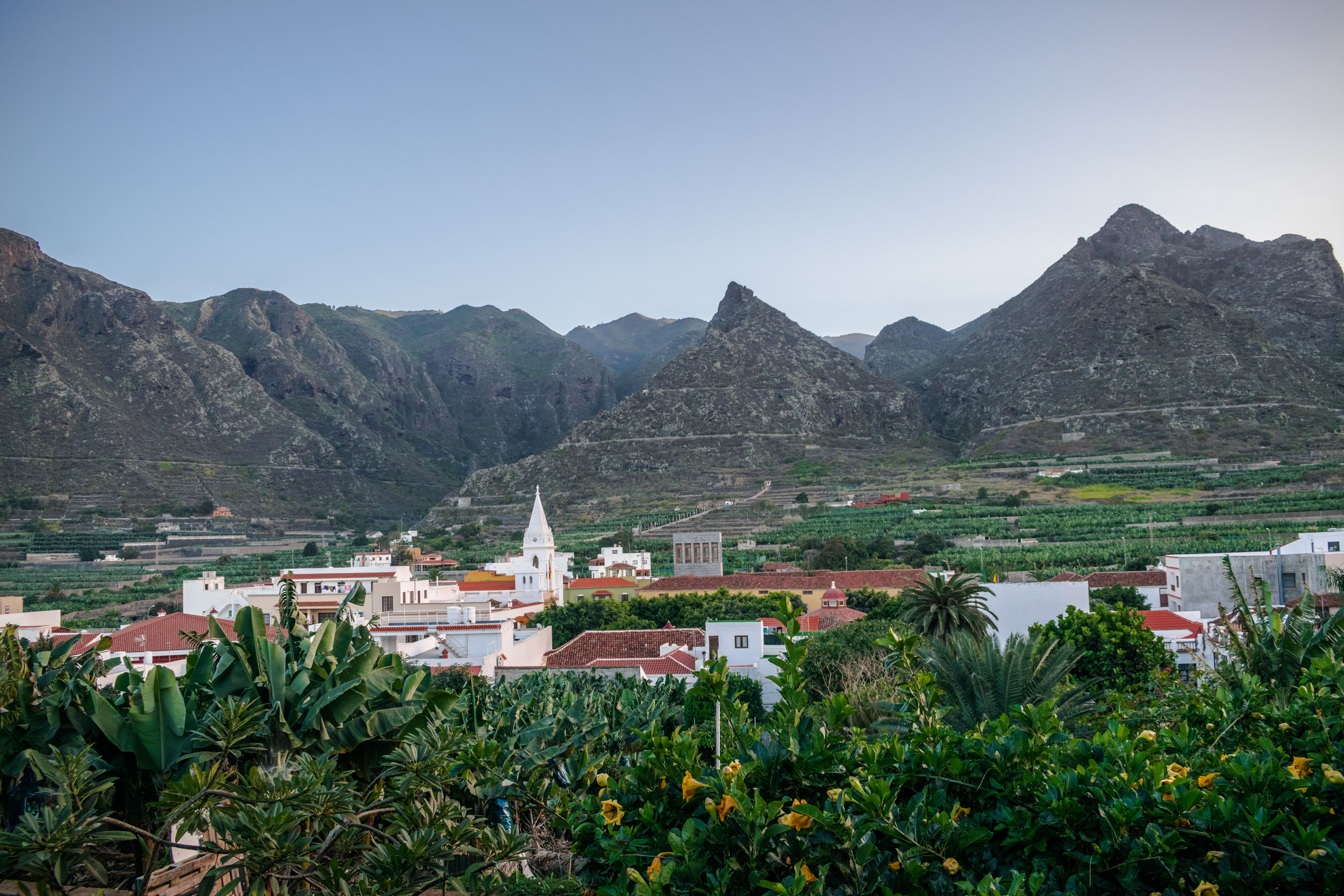 View of a valley town surrounded by terraced fields and backed by rocky mountains in soft sunset light.