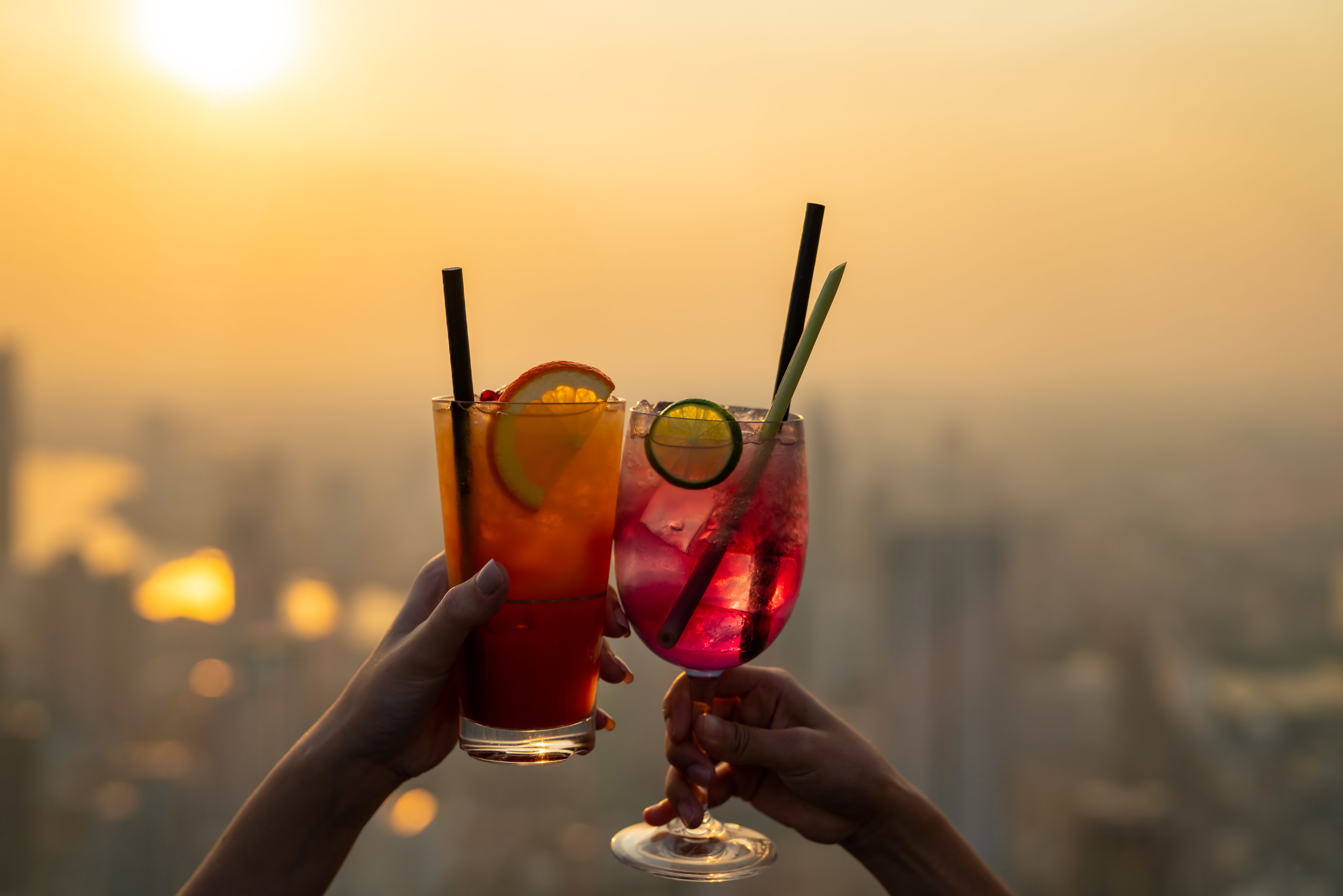Two people cheers-ing colourful cocktails together at a rooftop bar with a hazy summer background.