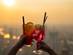 Two people cheers-ing colourful cocktails together at a rooftop bar with a hazy summer background.