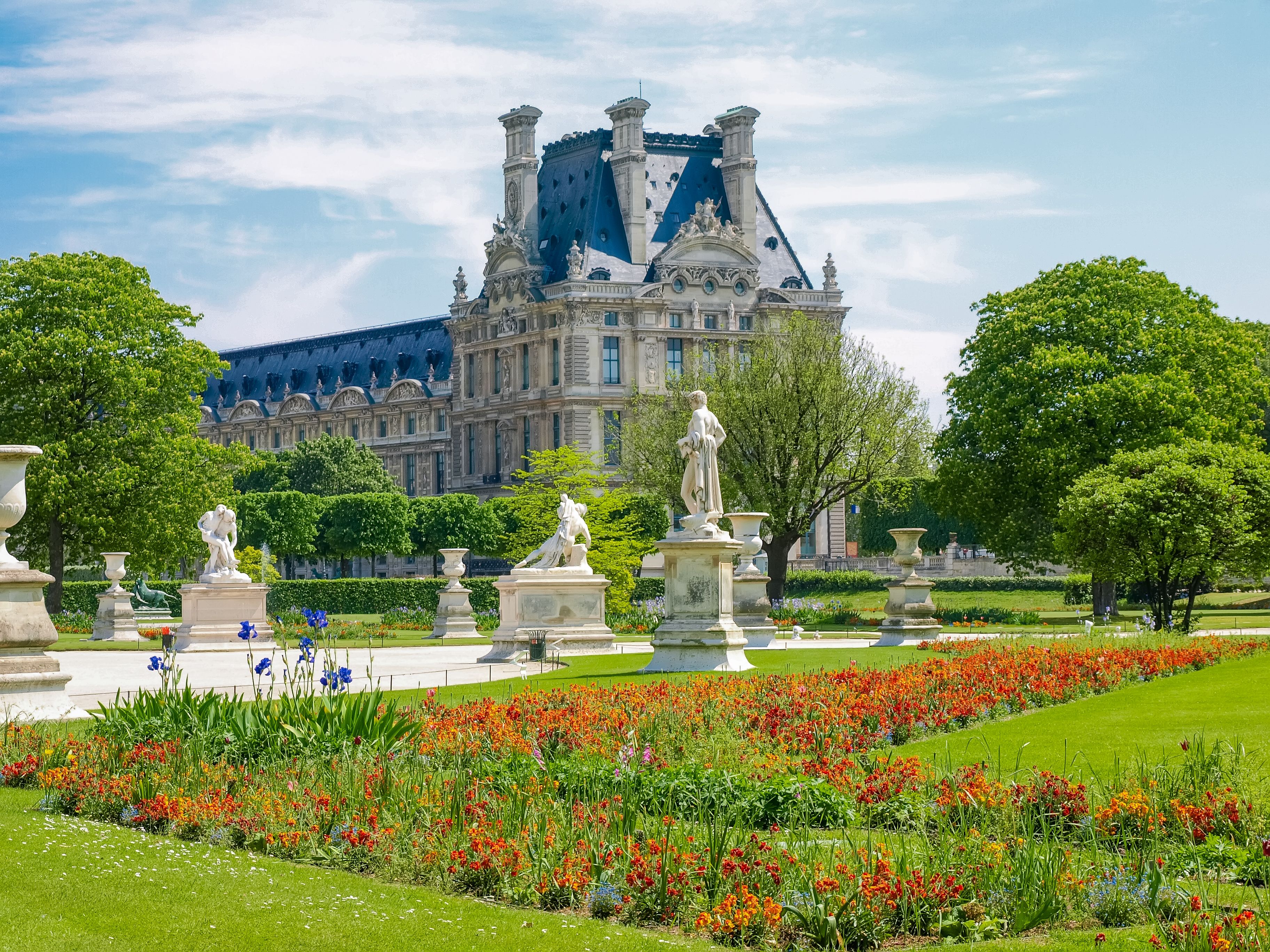 A view of the Tuileries Garden in Paris with lush green grass, bright flower beds and statue figures lining a path