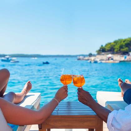 View of two people toasting an orange cocktail on a beach deck over the ocean