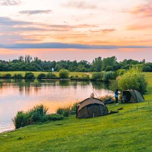 A view of fishing tents by the side of a lake in the UK countryside surrounded by green grass and with the sun setting