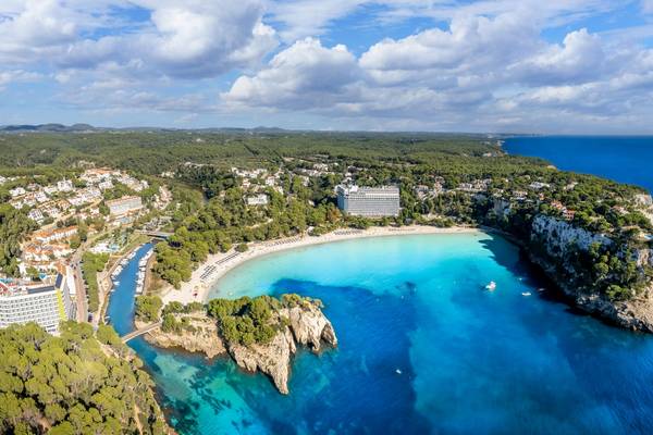 An aerial view of Cala Galdana beach in Menorca, Balearic Islands with beachfront hotels, an azure-coloured bay and a landscape of the island's green interior in the background