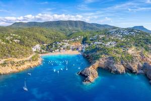 An aerial view of Cala Vadella bay and beach resort in Ibiza, Spain, with a rocky seashore, green mountainous land and blue sea