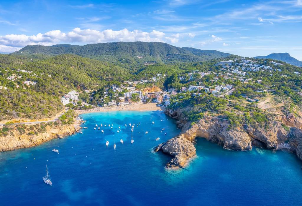 An aerial view of Cala Vadella bay and beach resort in Ibiza, Spain, with a rocky seashore, green mountainous land and blue sea