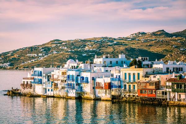 A view looking across the water of Little Venice in Mykonos town, Greece with white-washed houses and green hills in the background