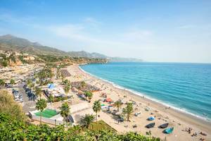 A view of Nerja beach and coastline in Costa del Sol, Spain with beach resorts and palm trees lining the coast and mountains in the background