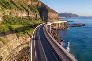 Aerial view of a single car driving along a bridge that runs alongside a cliff face and the Tasman Sea