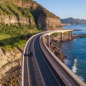 Aerial view of a single car driving along a bridge that runs alongside a cliff face and the Tasman Sea
