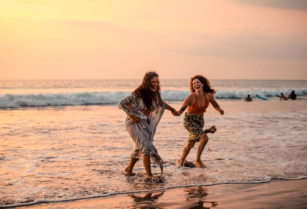 A wide shot of two female friends running out of the sea at Kuta beach in Bali, Indonesia at dusk while holding hands and laughing with each other.