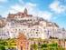A front-on view of a whitewashed town perched on a clifftop with a grand cathedral overlooking it all.