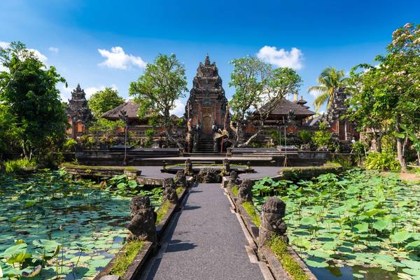 A path with lotus ponds on either side leads to an ornate, stone Balinese temple with a towering, tiered gate as the threshold to the temple