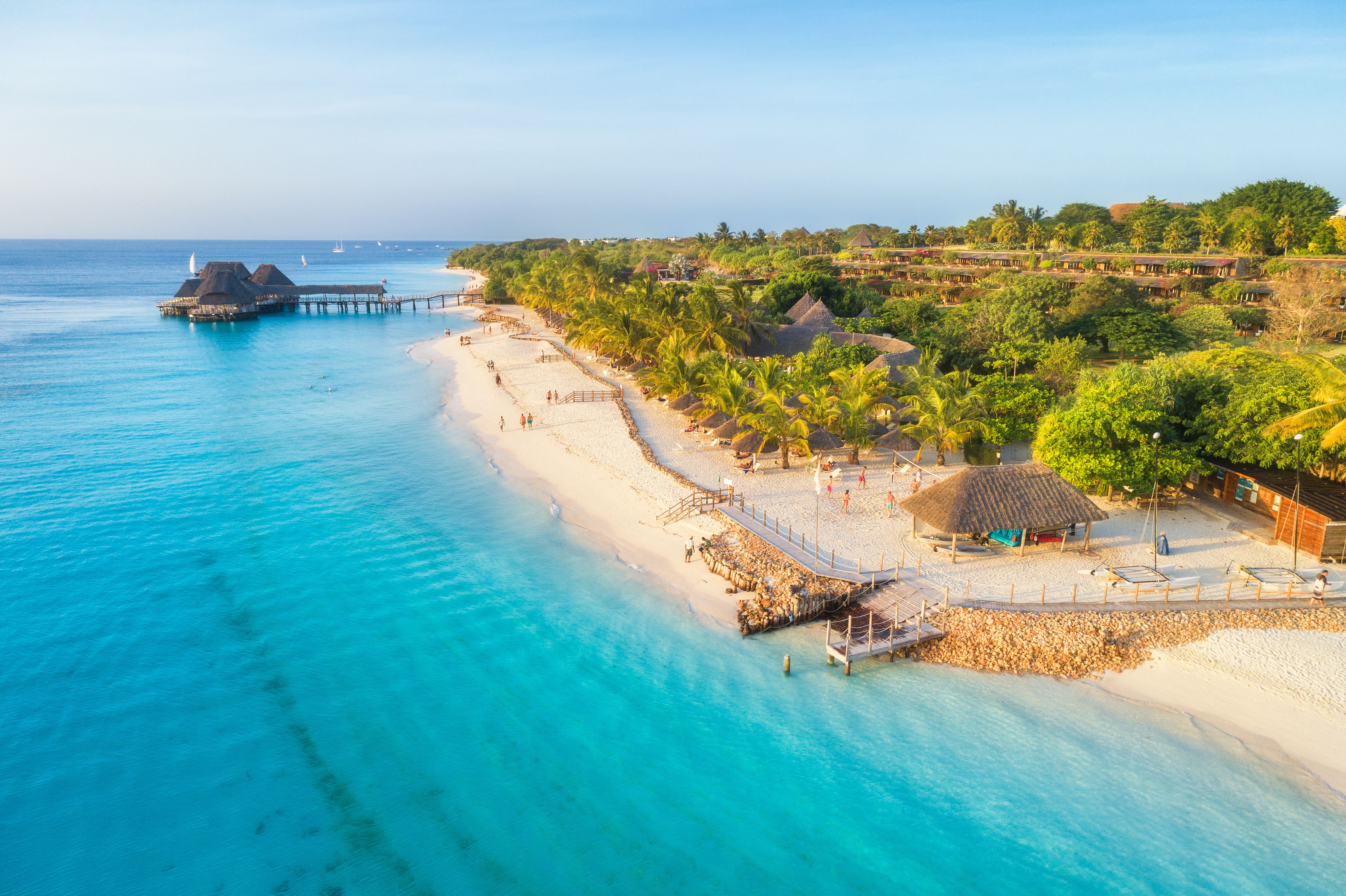 View of a sandy beach lined with palm trees and lapped by turquoise waters, with over-water villas in the distance.