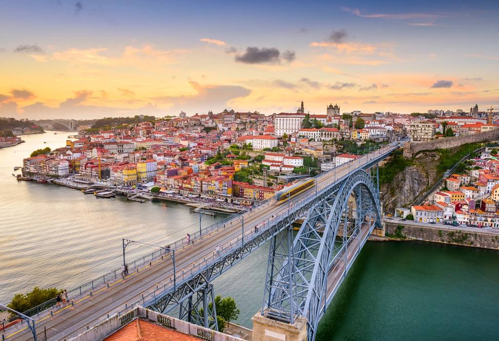 Aerial view of Porto from the Gaia side of the city, featuring the Dom Luis double-deck metal arch bridge crossing the Douro River and the city's skyline at sunset