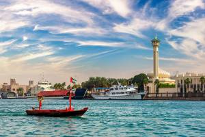 View of a red wooden boat with a triangular roof sailing on Dubai Creek with Dubai's old town in the background