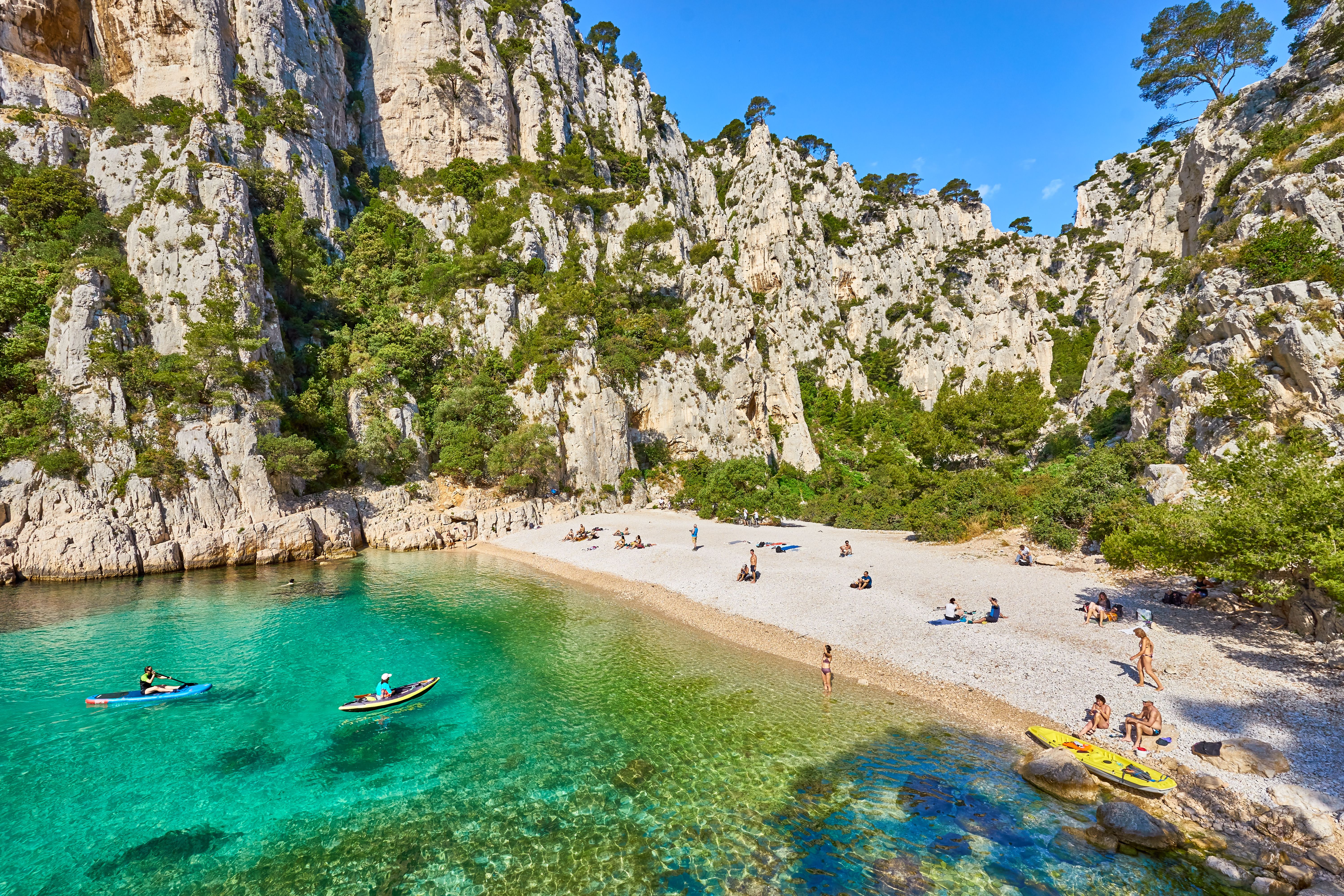 Aerial view of sunbathers and paddleboarders on a pebbly beach surrounded by dramatic limestone cliffs