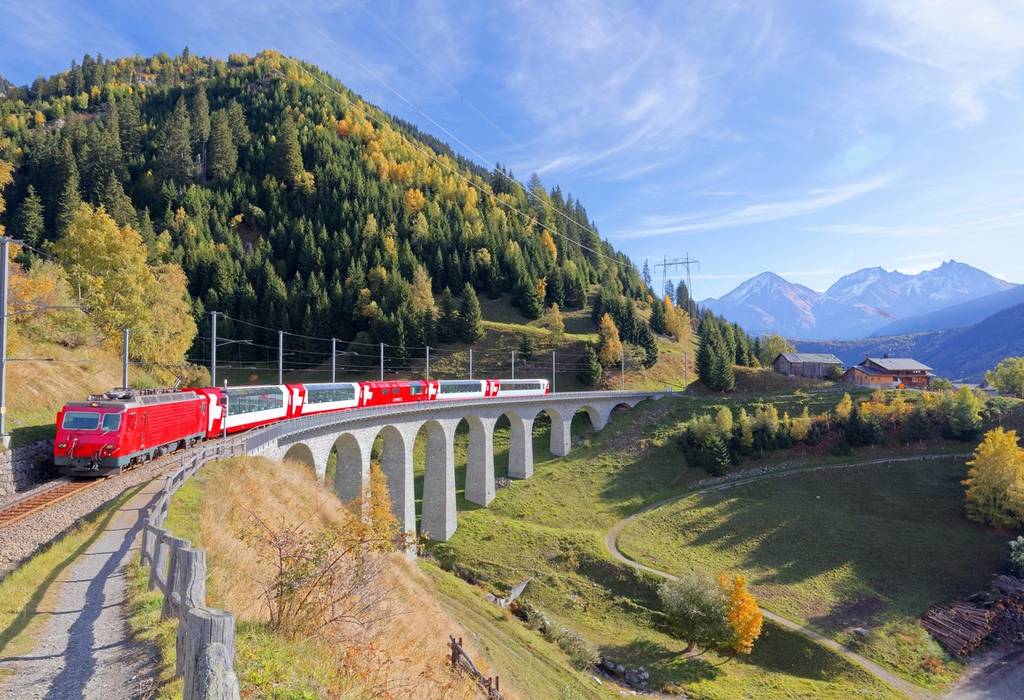 View of a red train with the Swiss flag on the side travelling across a viaduct on a backdrop of autumnal mountains