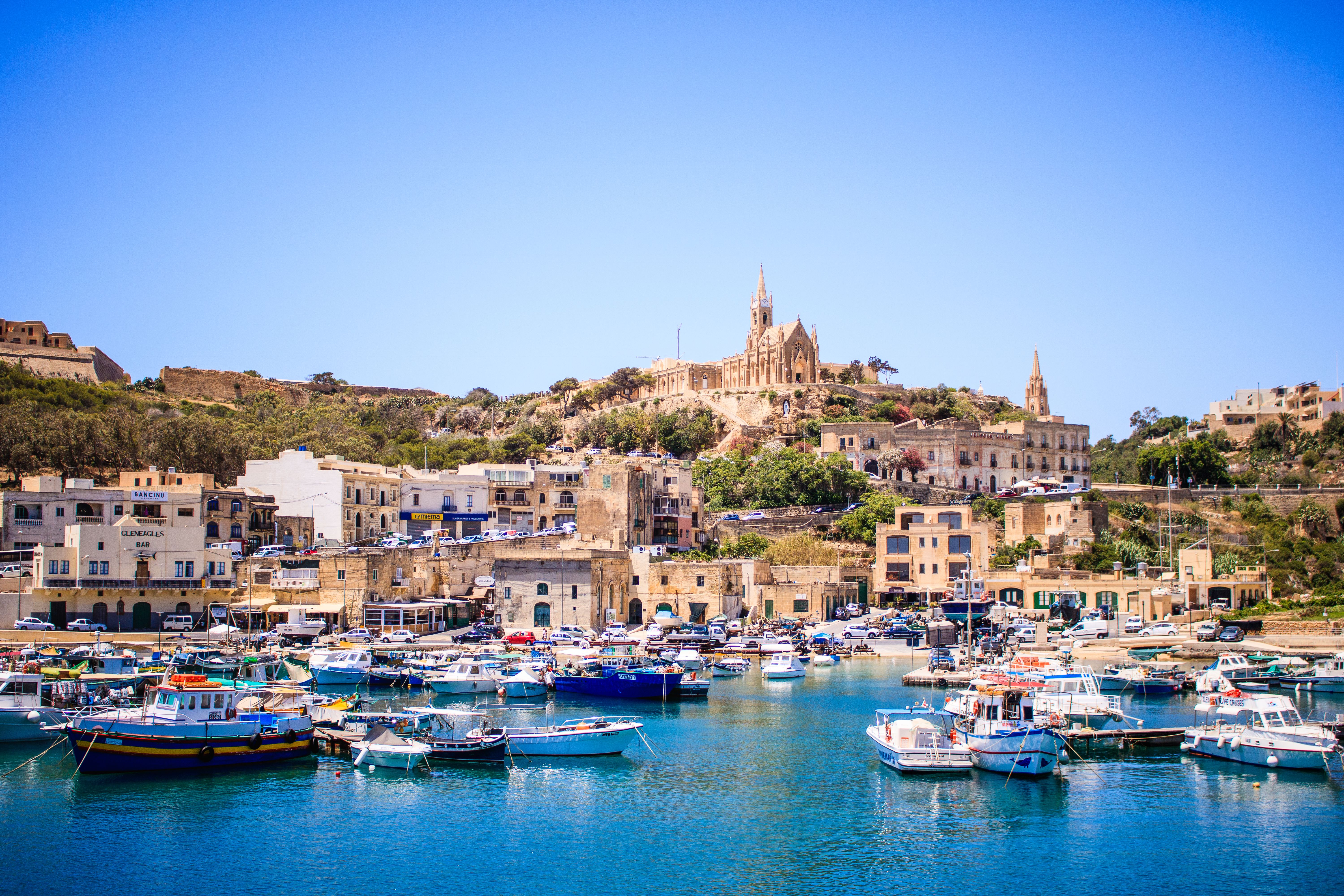 A harbour front view of the island of Gozo in Malta with buildings set into the hillside and colourful boats moored in the harbour