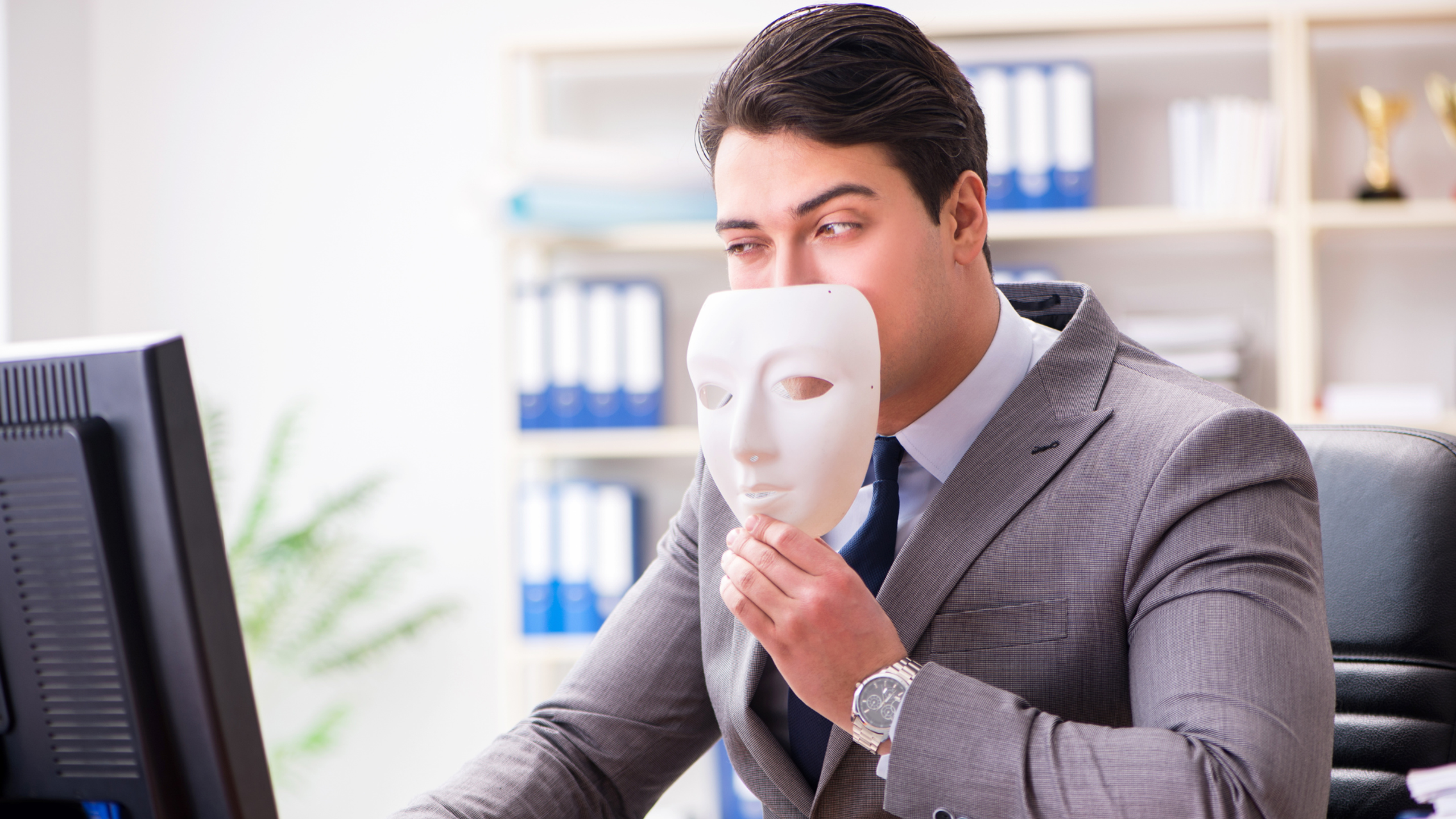 Photo of a businessperson at their desk holding a mask near their face as they look at their computer.