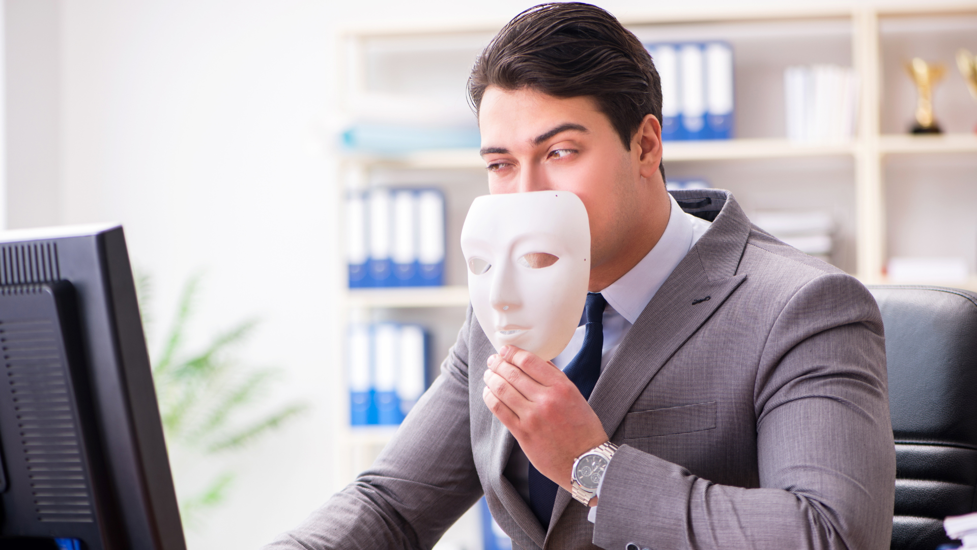 Photo of a businessperson at their desk holding a mask near their face as they look at their computer.