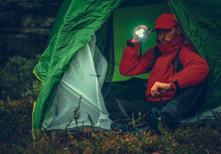 Caucasian Camper with Flashlight Inside His Tent During Rainy Evening While on Hike.