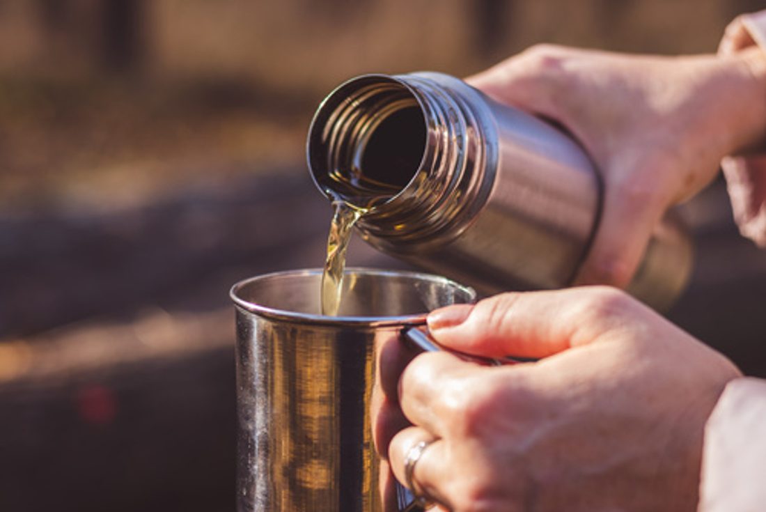 Woman pours hot tea out of thermos into metal cup in autumn fore
