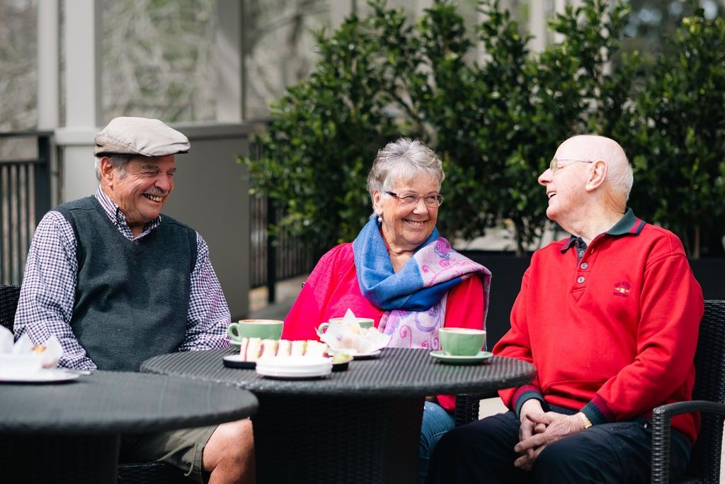 Group of smiling seniors enjoying coffee and cake outdoors at a New Zealand retirement village, sharing laughter and friendship on a sunny day.
