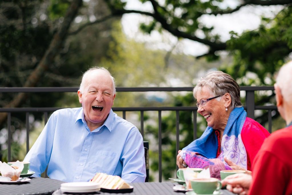 Group of happy seniors laughing together over coffee and cake outdoors on a sunny day, enjoying conversation at a retirement village café.