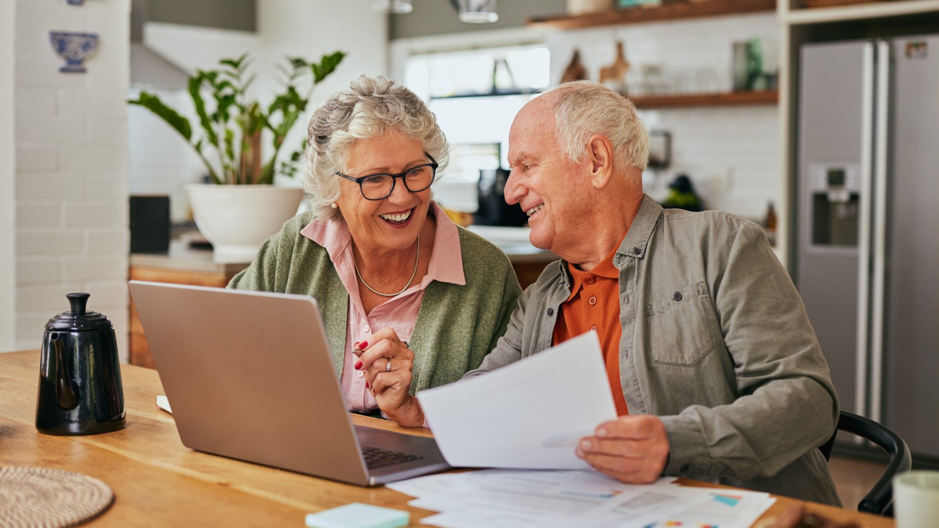 Senior couple smiling while reviewing documents and using a laptop at home, representing financial planning and retirement living with Metlifecare.