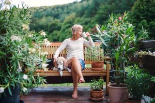 Older woman relaxing on a garden bench with her dog, enjoying a cup of tea surrounded by plants, reflecting peaceful retirement living.