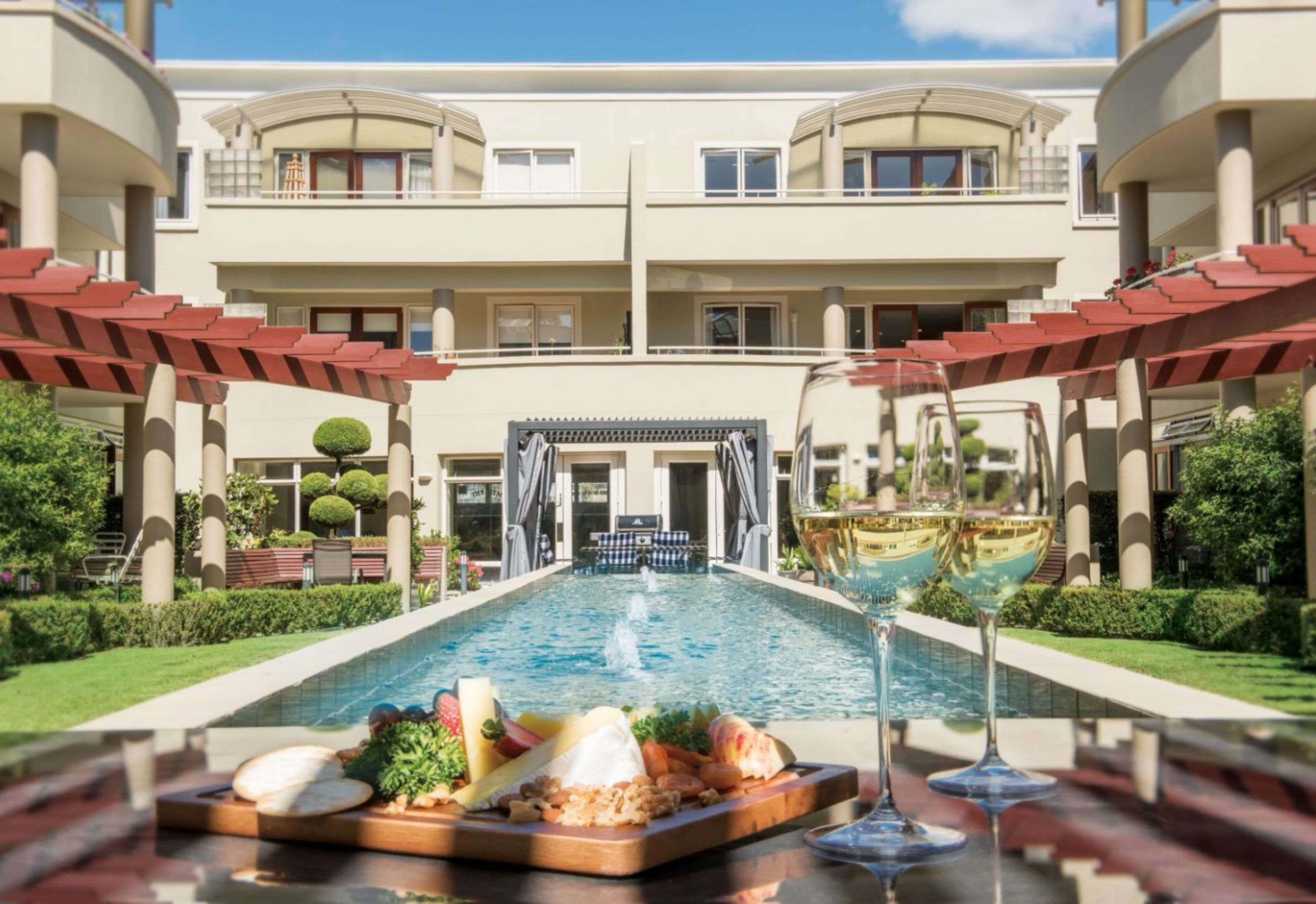 A table with a plate of food and two glasses of wine by a pool at the Avenues - A Metlifecare rest homes in the Bay of Plenty.