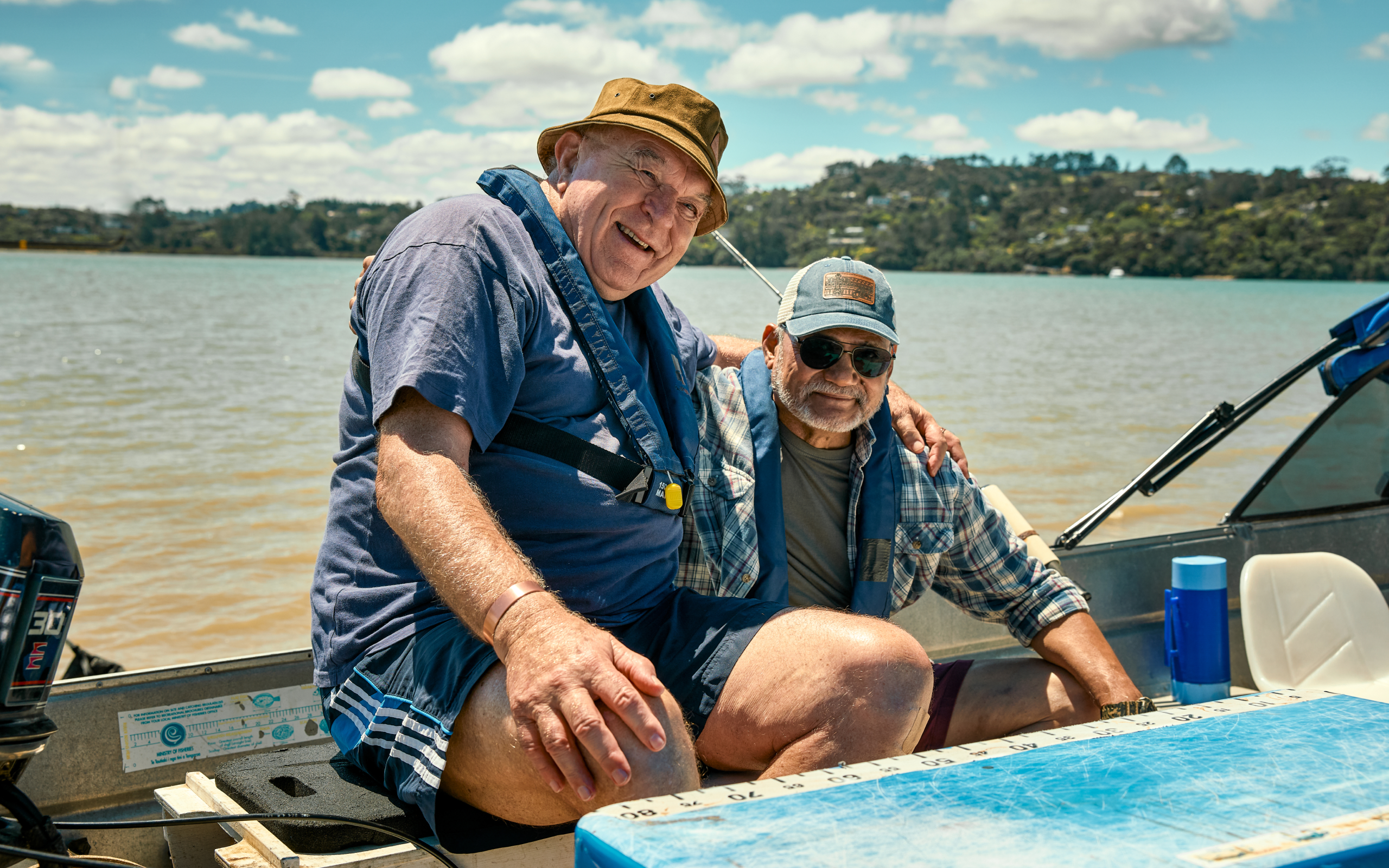 Two older men enjoying a fishing trip on a boat in New Zealand, relaxing on the water with scenic coastal hills in the background.