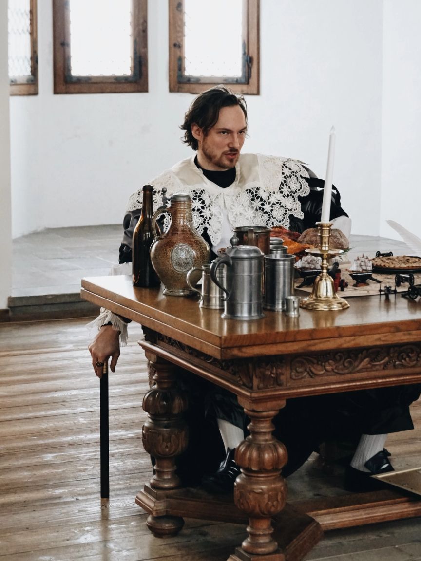 Skuespiller i historisk dragt sidder ved bord.