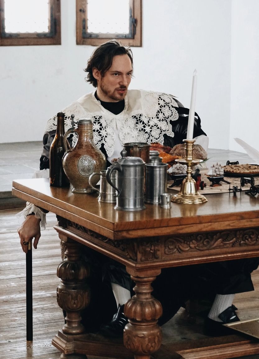 Skuespiller i historisk dragt sidder ved bord.