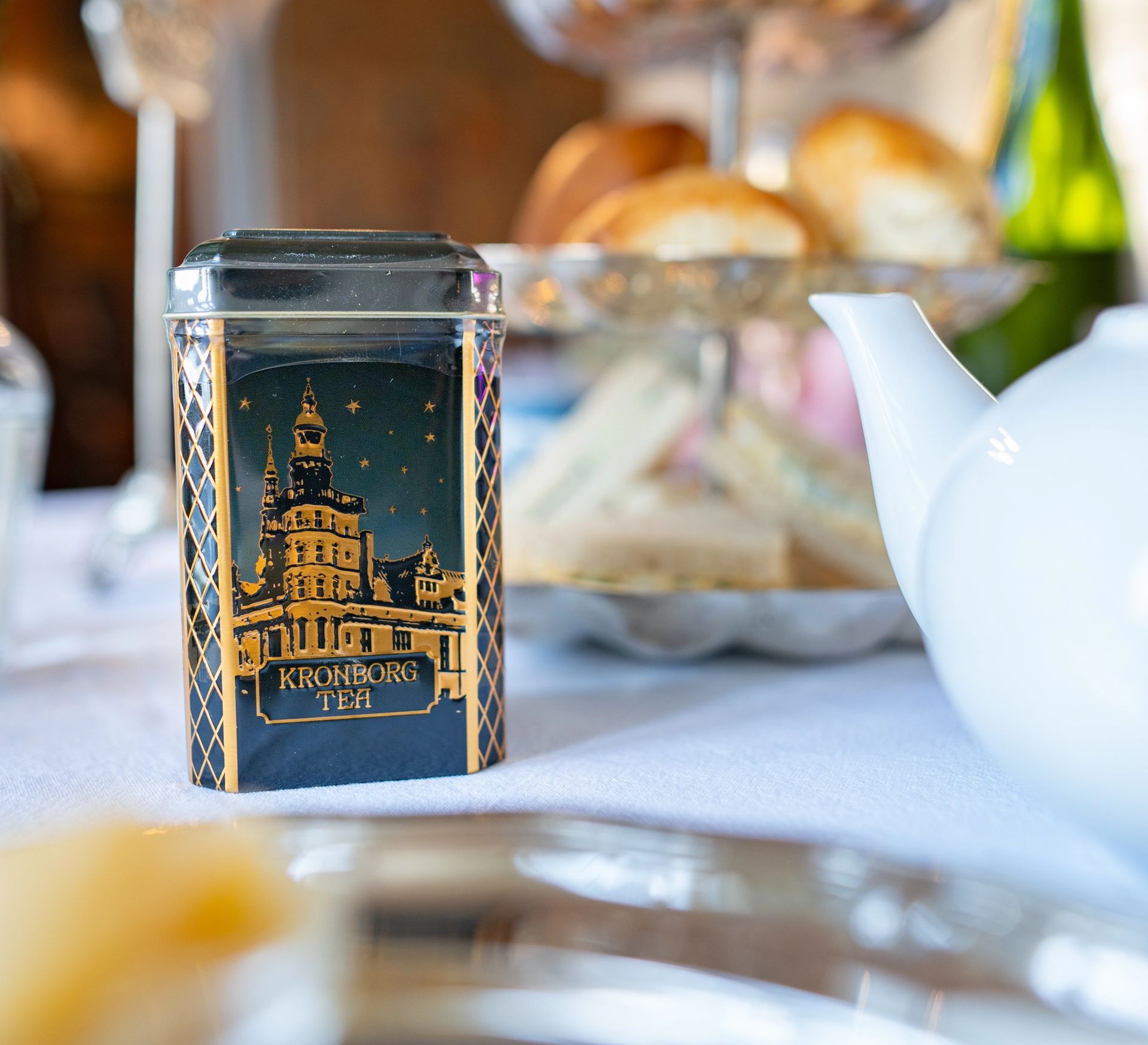 Kronborg-tedåse med slottets motiv og teksten 'Kronborg Teh' på et afternoon tea-bord – souvenirprodukt som del af oplevelsen på slottet