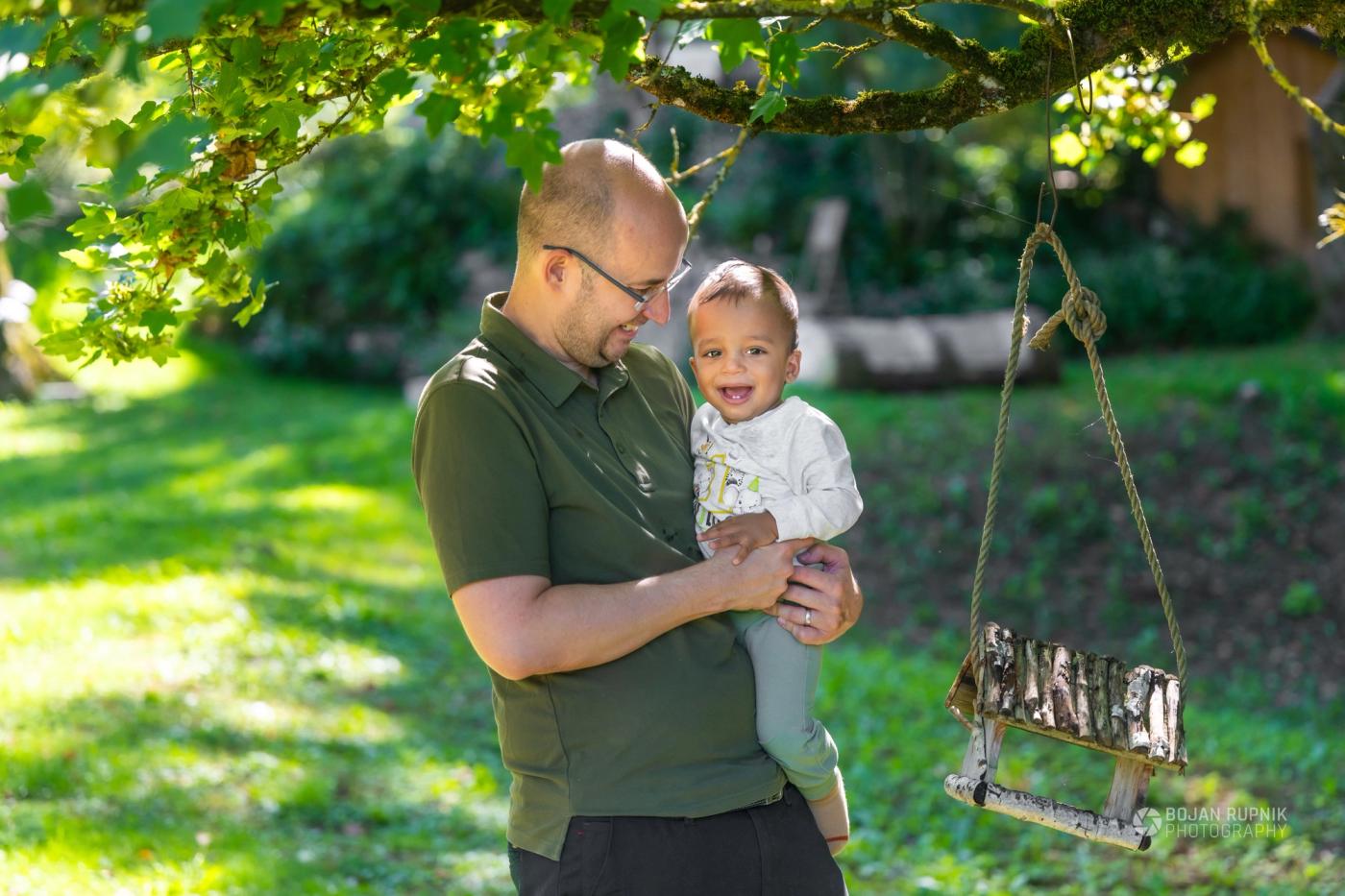 Nasmejan oče drži malega sina pod drevesom ob leseni gugalnici