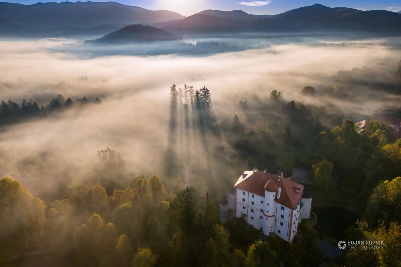 Bel grad z rdečo streho v gozdu, prepredenem z jutranjo meglo in sončnimi žarki, posnetek iz zraka