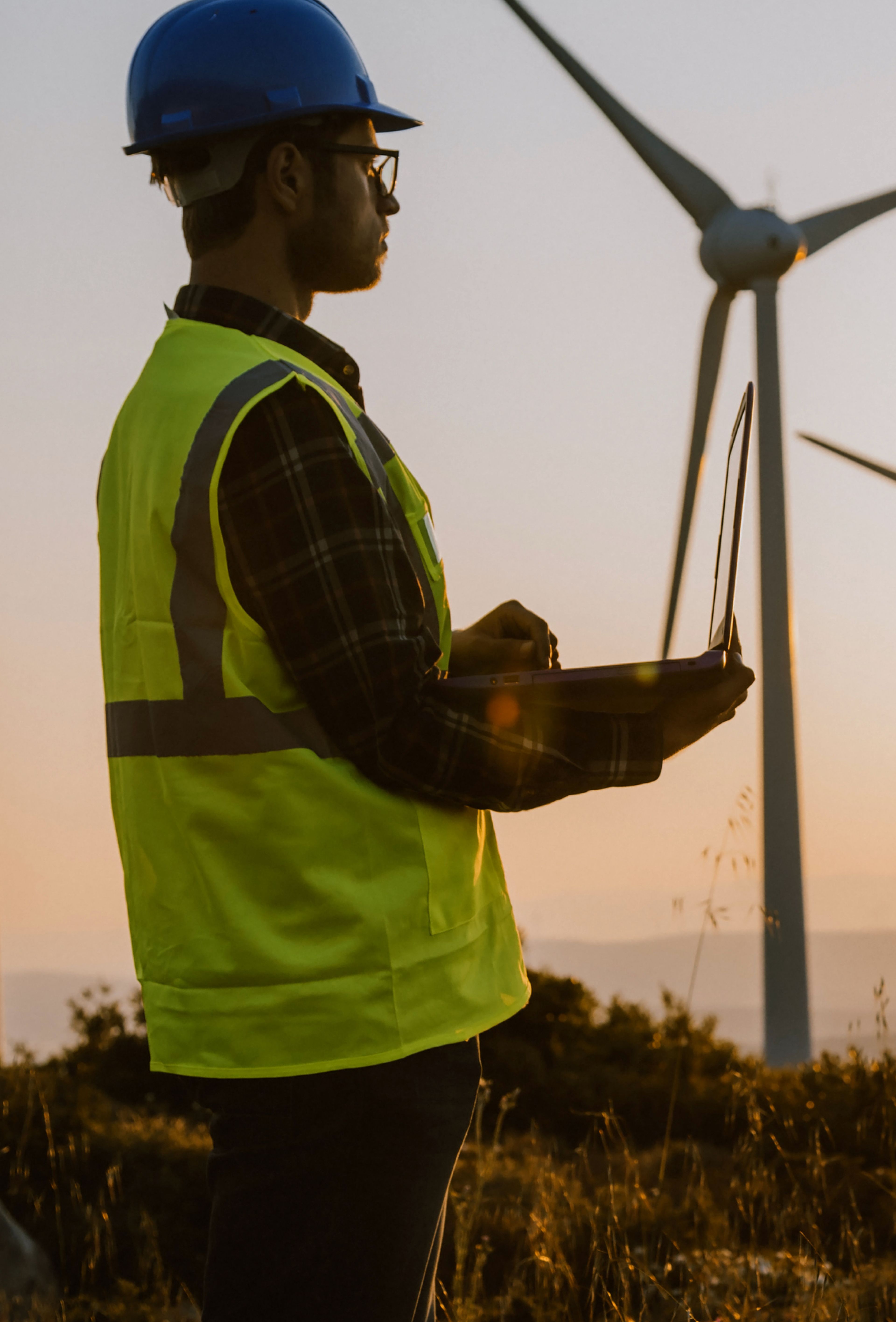 African American man wearing black hard hat and yellow safety vest holding a clipboard against a sunset horizon lined with eight wind turbines