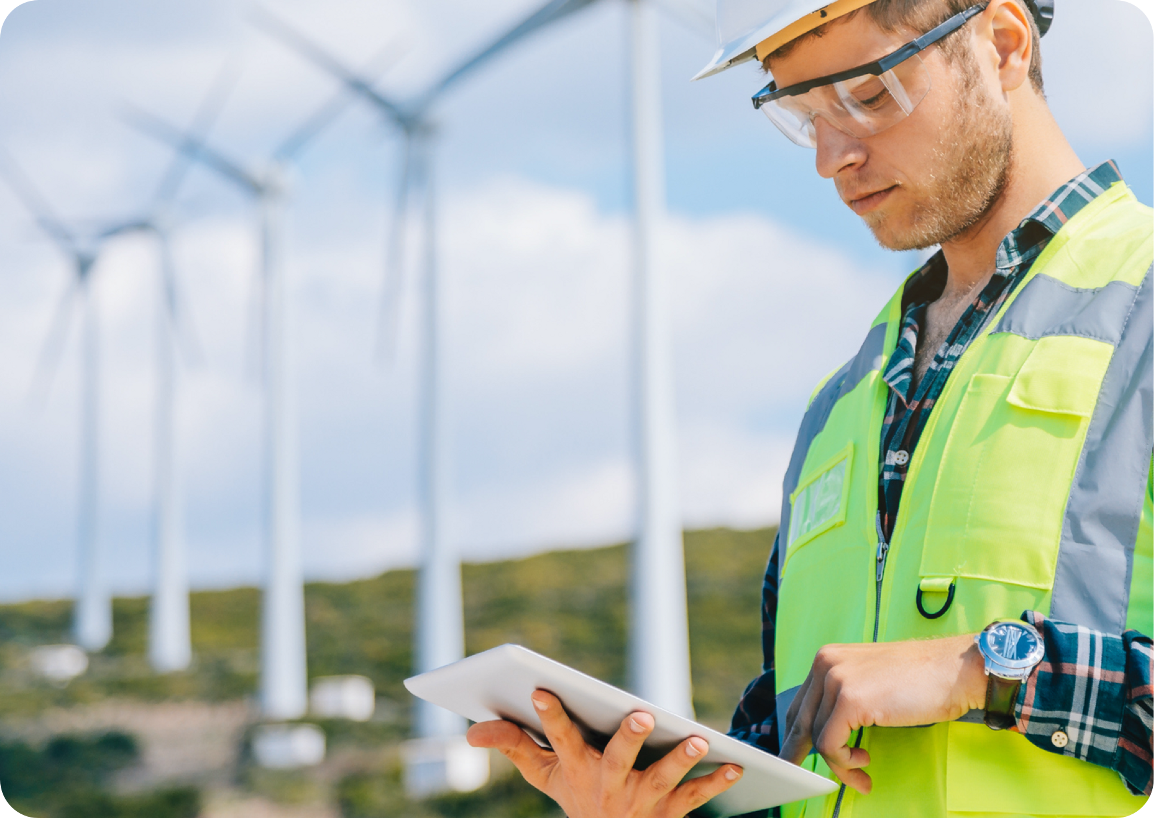 Picture of Caucasian man wearing safety glasses and yellow safety vest typing on an iPad and three wind turbines spinning in the background 