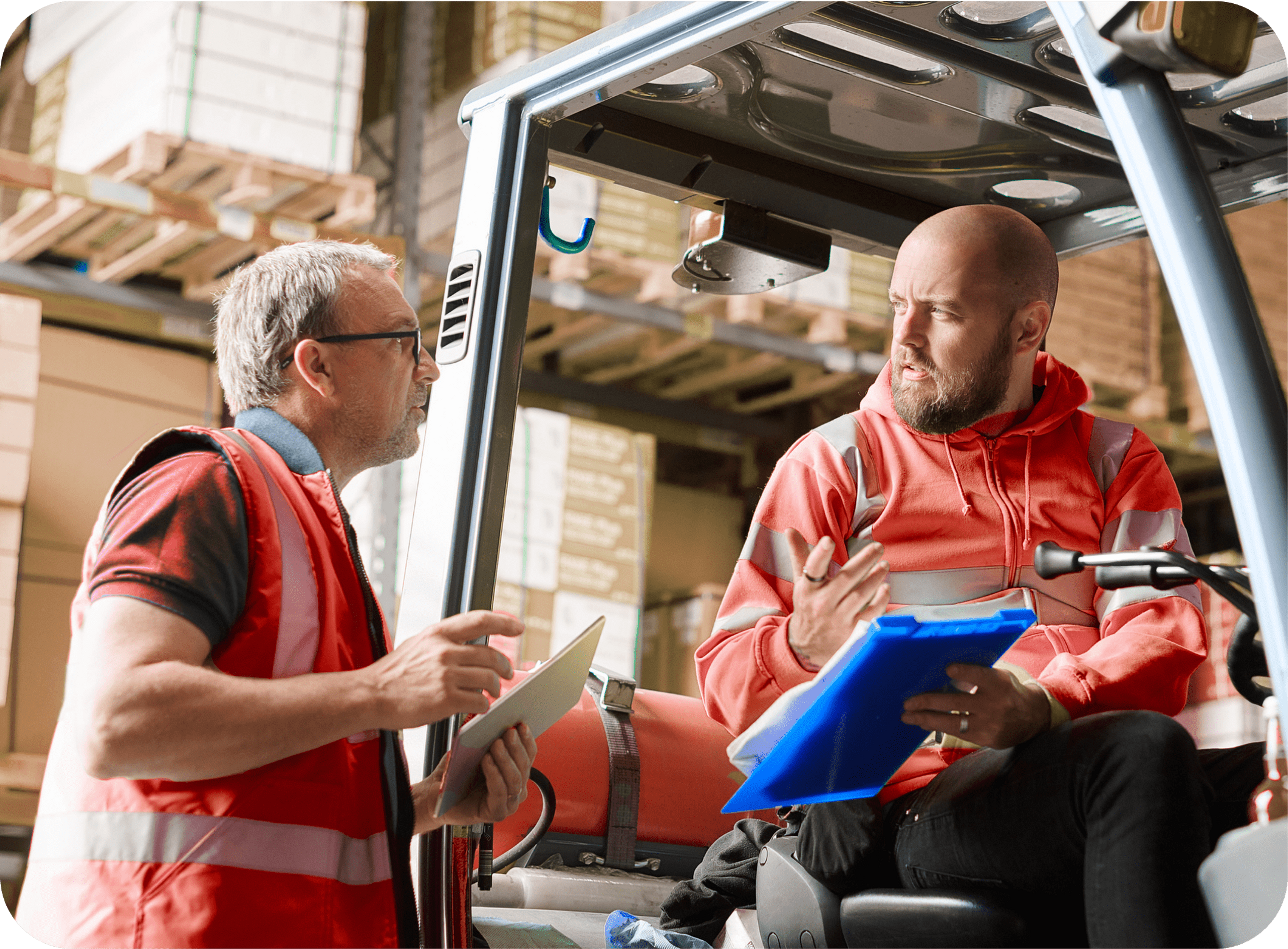 Two Caucasian men in red safety vests talking to each other in a warehouse setting