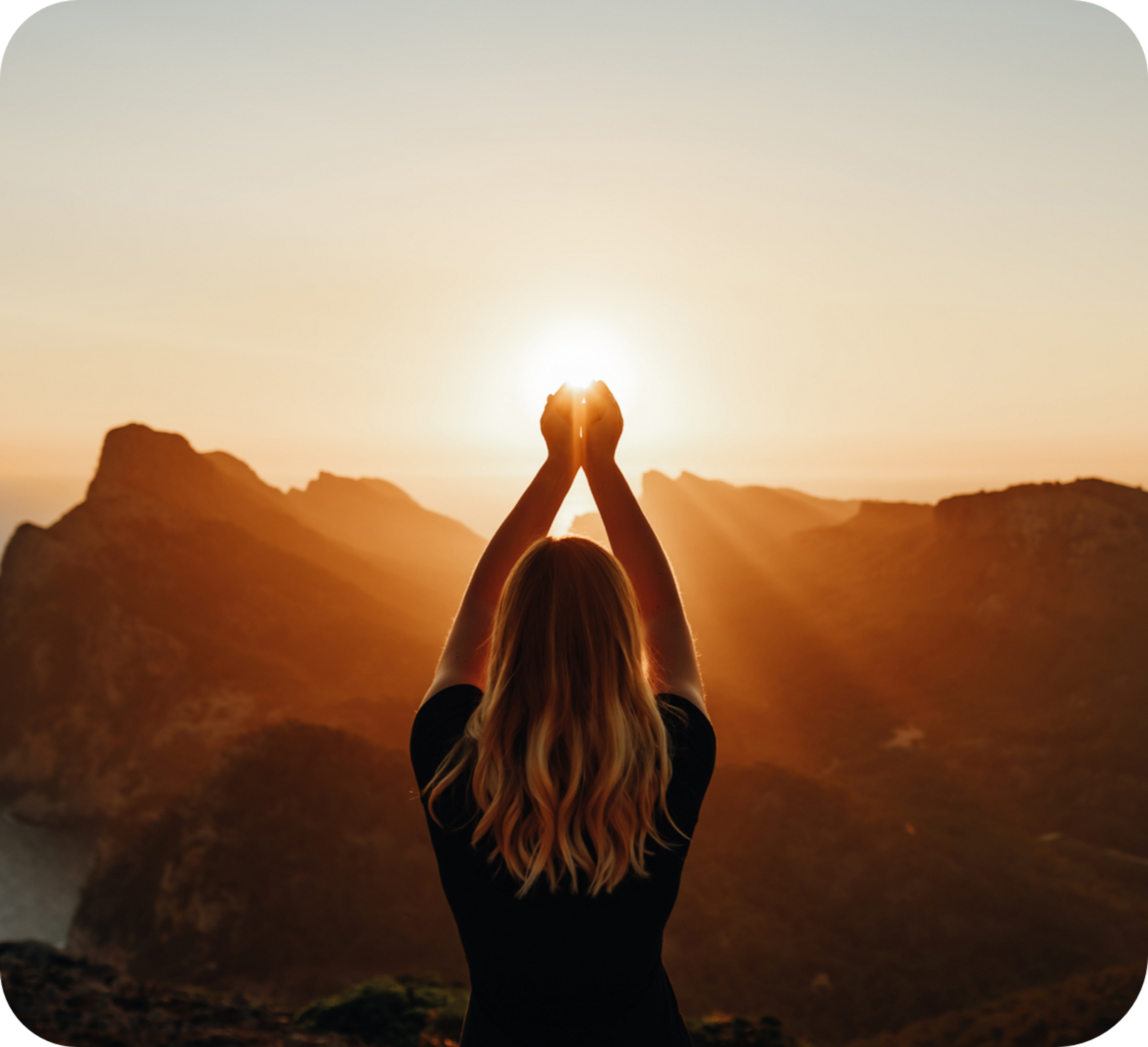 Photo d'une femme de dos entourée de montagnes, les mains réunies face à un coucher de soleil