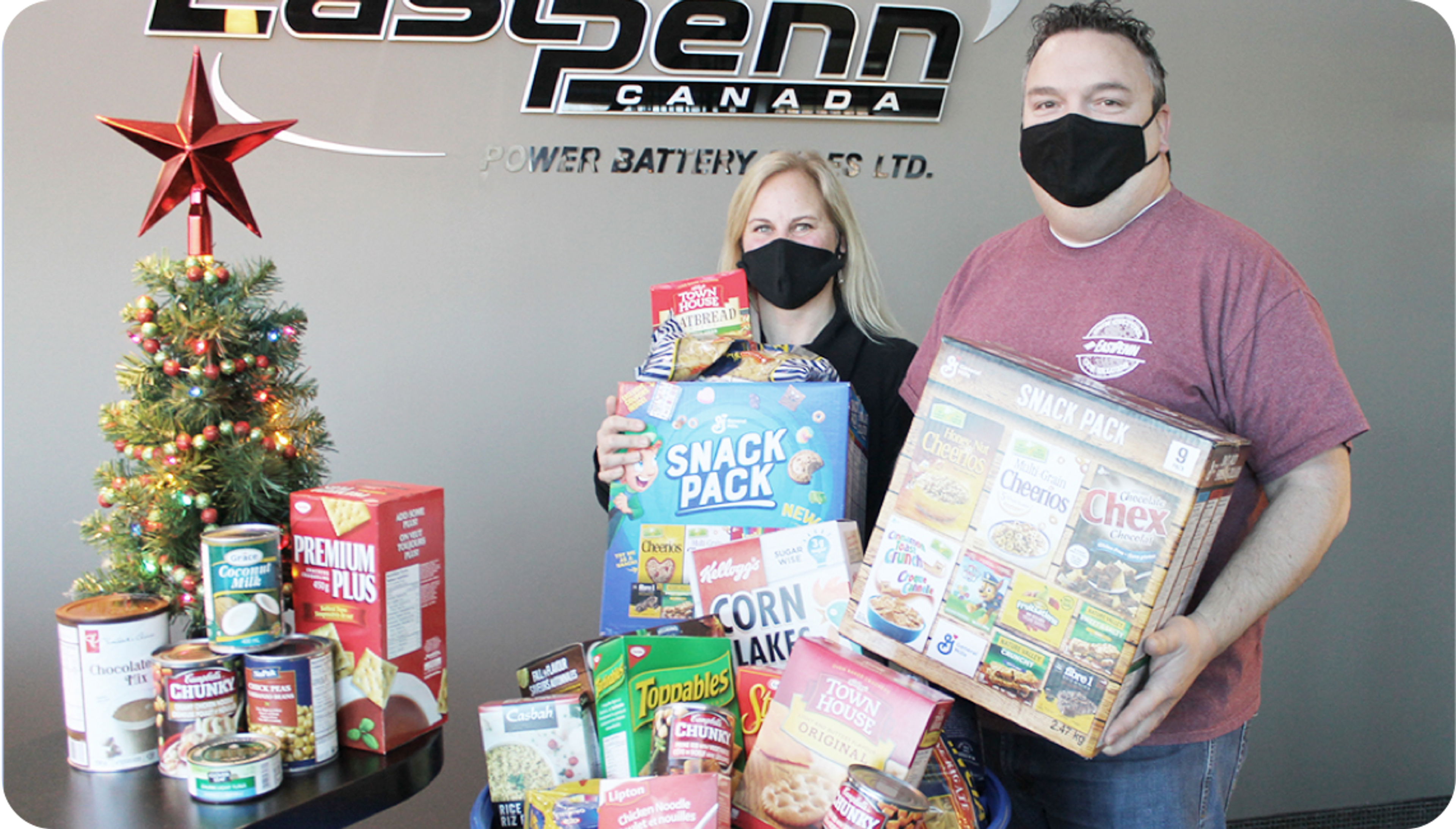 Image of a masked man and woman holding food donations next to a tabletop Christmas tree.