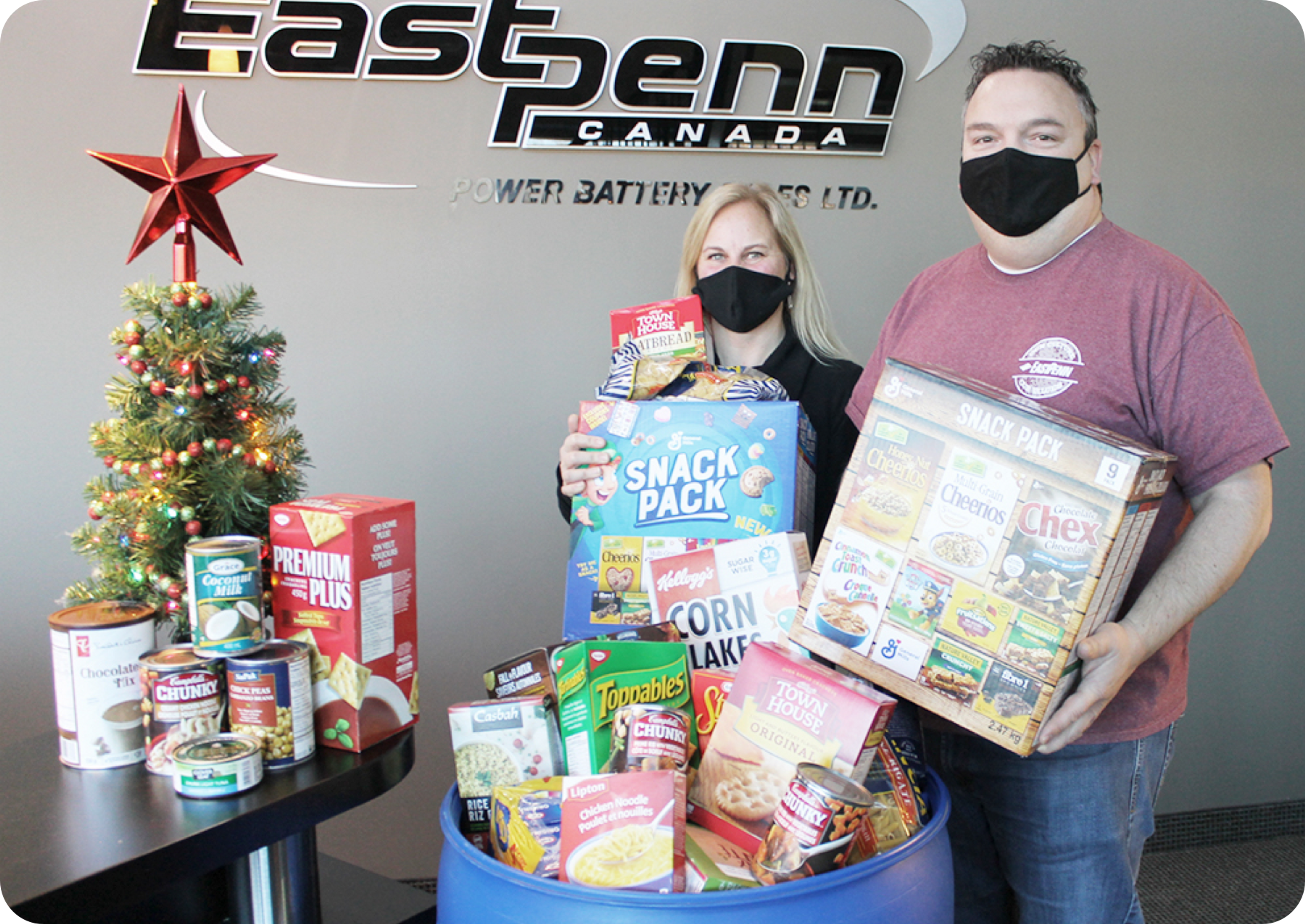 Image of a masked man and woman holding food donations next to a tabletop Christmas tree.