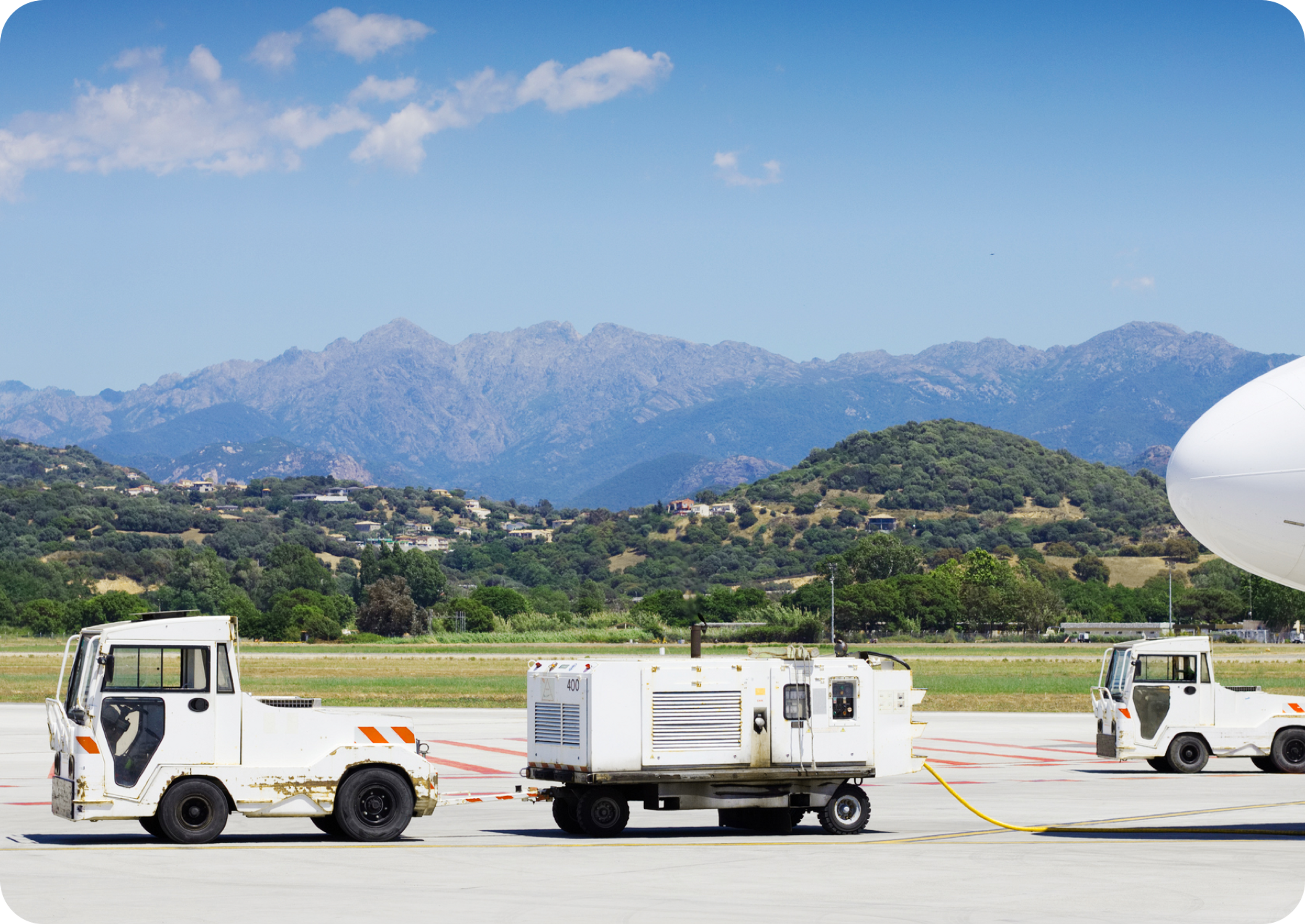 Pictire of white truck and white battery trailer at an airport runway with trees and mountains in the horizon