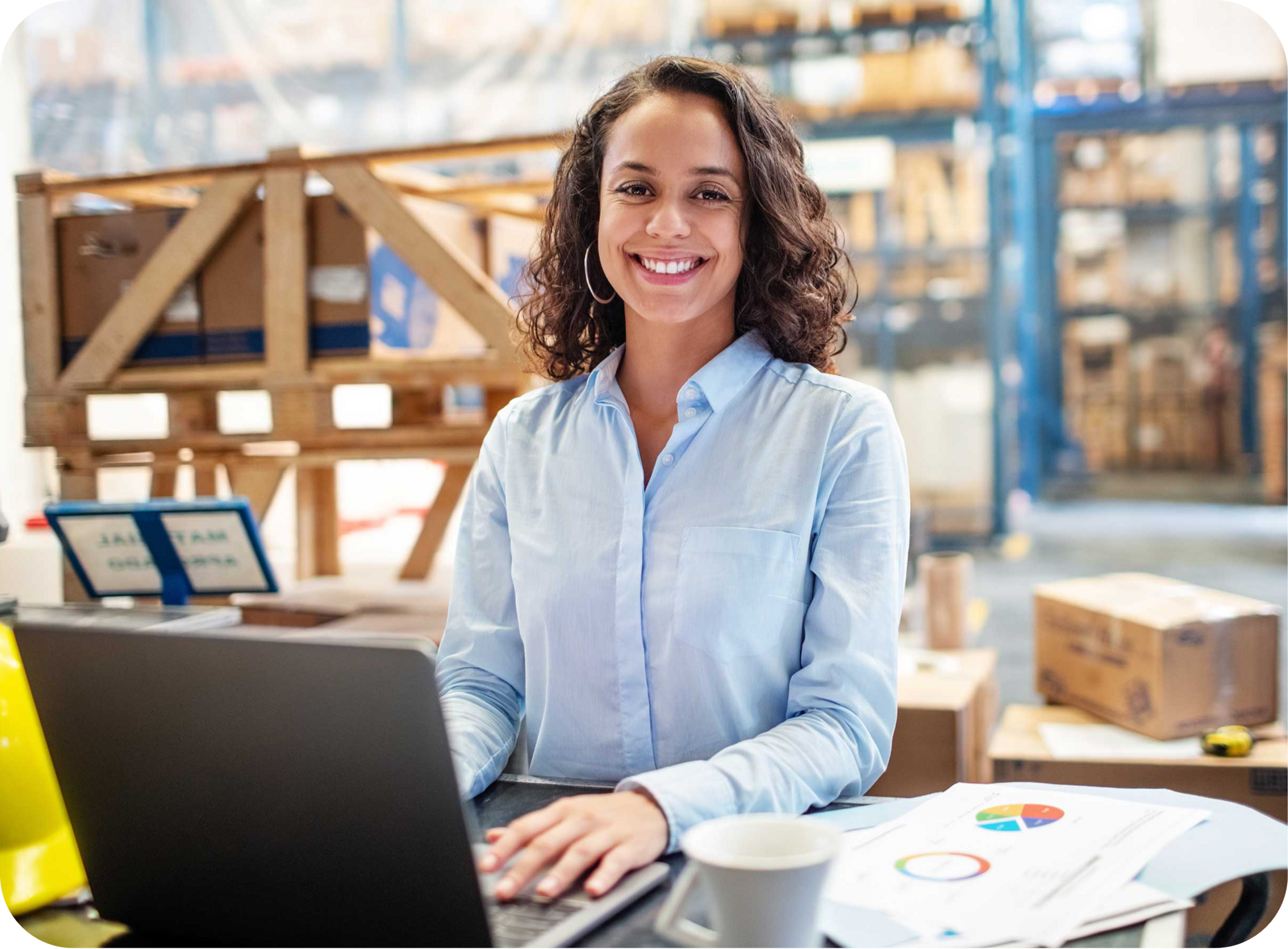 Picture  of Hispanic woman wearing a blue dress shirt, smiling and using a laptop on a desk with a coffee cup, graph inside a warehouse